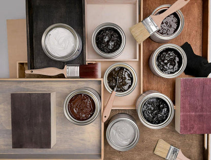An overhead flat-lay of multiple open metal paint cans containing different paint colors, including white, black, brown, and gray, arranged on wooden trays with paintbrushes and wood blocks.