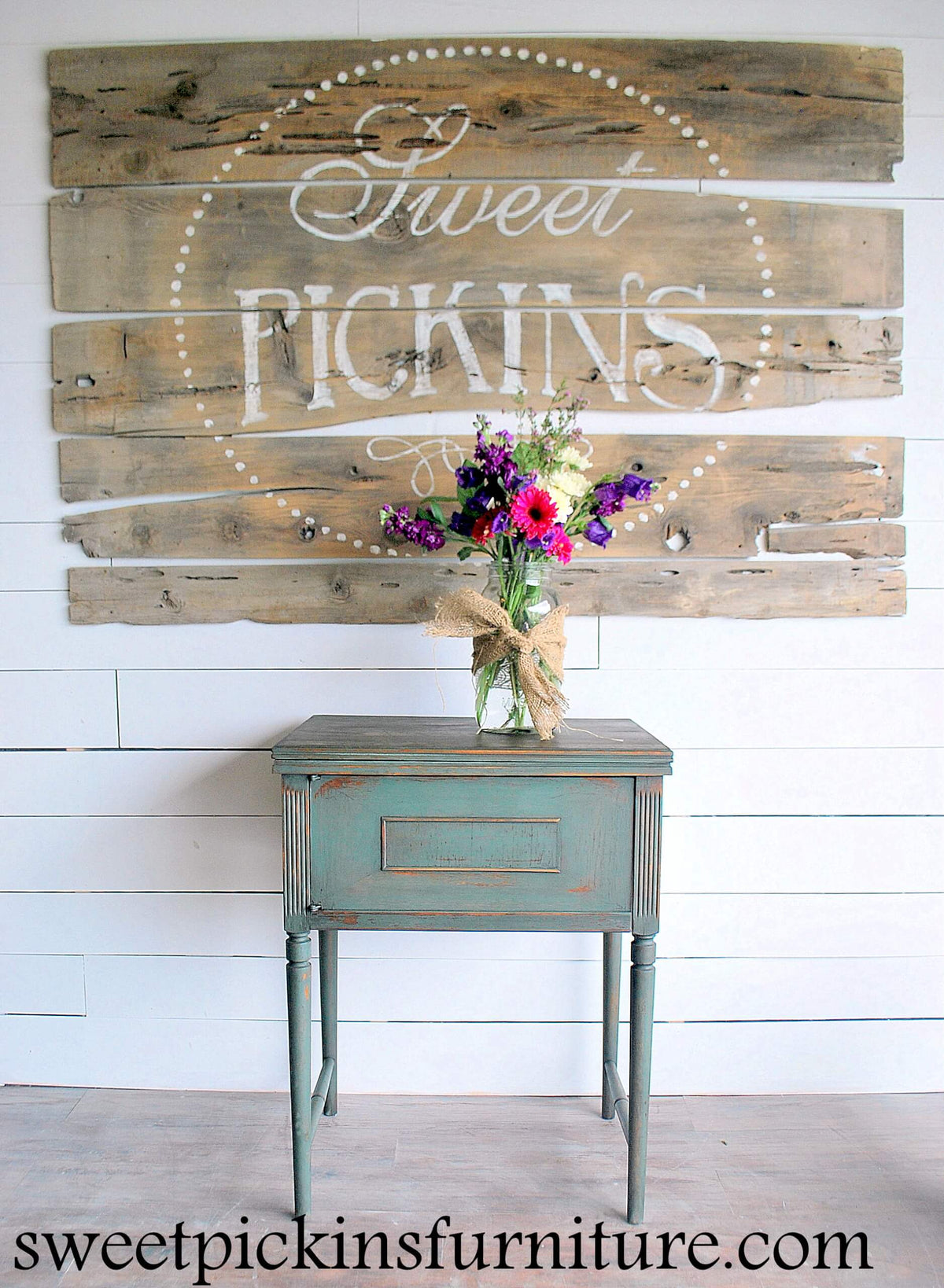 Wooden sign with 'Sweet Pickins' on a white wall with a small table and flowers.