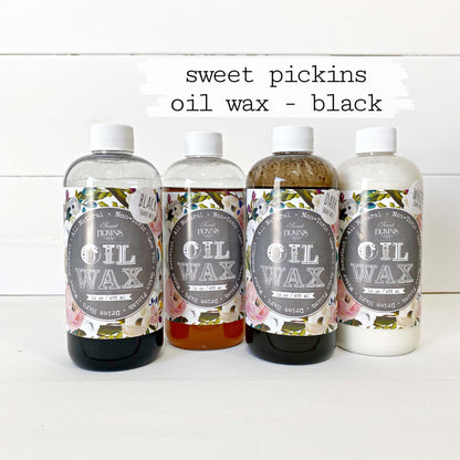 Four bottles of Sweet Pickins Natural Oil Wax arranged in a row on a white surface against a white wood plank background. The top of the image has a white brush-stroke style banner with the text 'sweet pickins oil wax - black'.