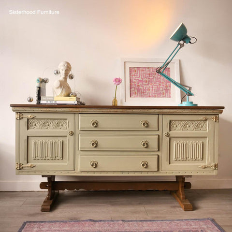 A vintage-style sideboard or dresser in a light sage green finish with a dark wood top and legs, styled in a room setting with a teal desk lamp, a bust sculpture, and framed art.