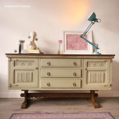 A vintage-style sideboard or dresser in a light sage green finish with a dark wood top and legs, styled in a room setting with a teal desk lamp, a bust sculpture, and framed art.