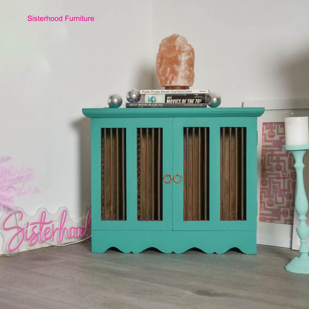A turquoise painted wooden cabinet with vertical slatted glass doors and gold ring handles, styled in a room setting with a salt lamp, books, and decorative items on top.