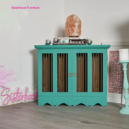 A turquoise painted wooden cabinet with vertical slatted glass doors and gold ring handles, styled in a room setting with a salt lamp, books, and decorative items on top.