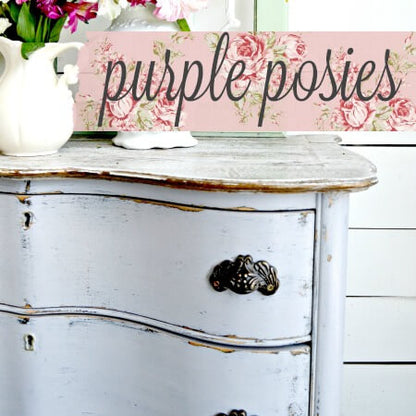 A rectangular wooden sign with the words 'purple posies' in black script over a pink floral background, displayed on top of a distressed white dresser with a white pitcher of flowers nearby.
