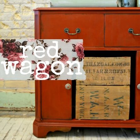 A red painted wooden cabinet or dresser with two upper drawers, one lower cabinet door, and an open lower section holding vintage wooden boxes. A floral banner overlay reads 'red wagon' across the front.
