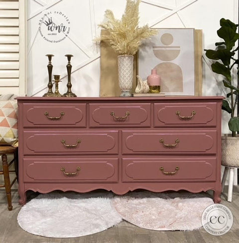 A dusty rose six-drawer dresser with brass handles, styled in a home interior setting with decorative items on top, a plant, and two round shag rugs on the floor.