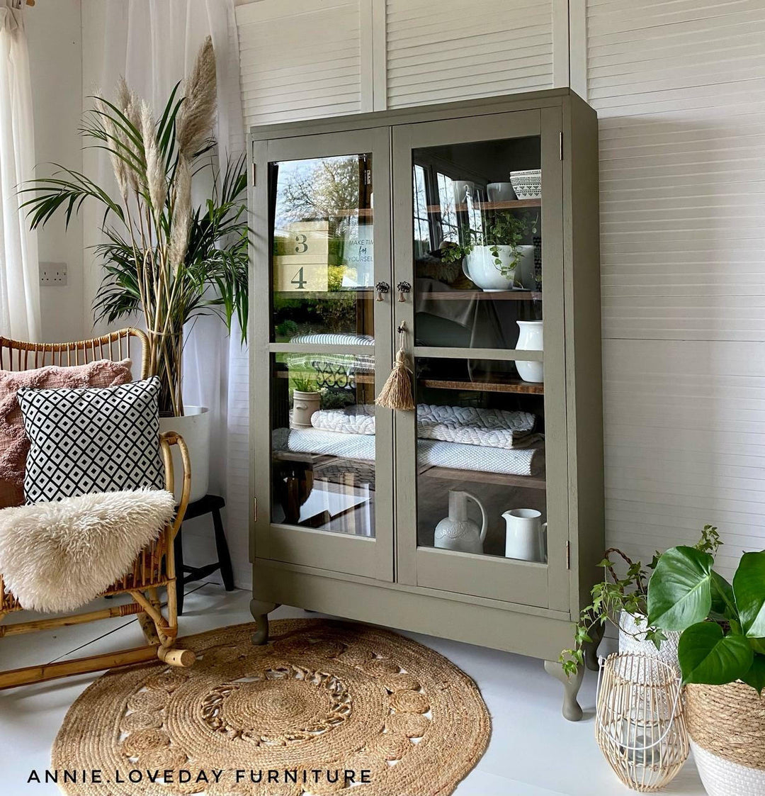 Green glass-fronted cabinet in a room with a chair, rug, and plants.