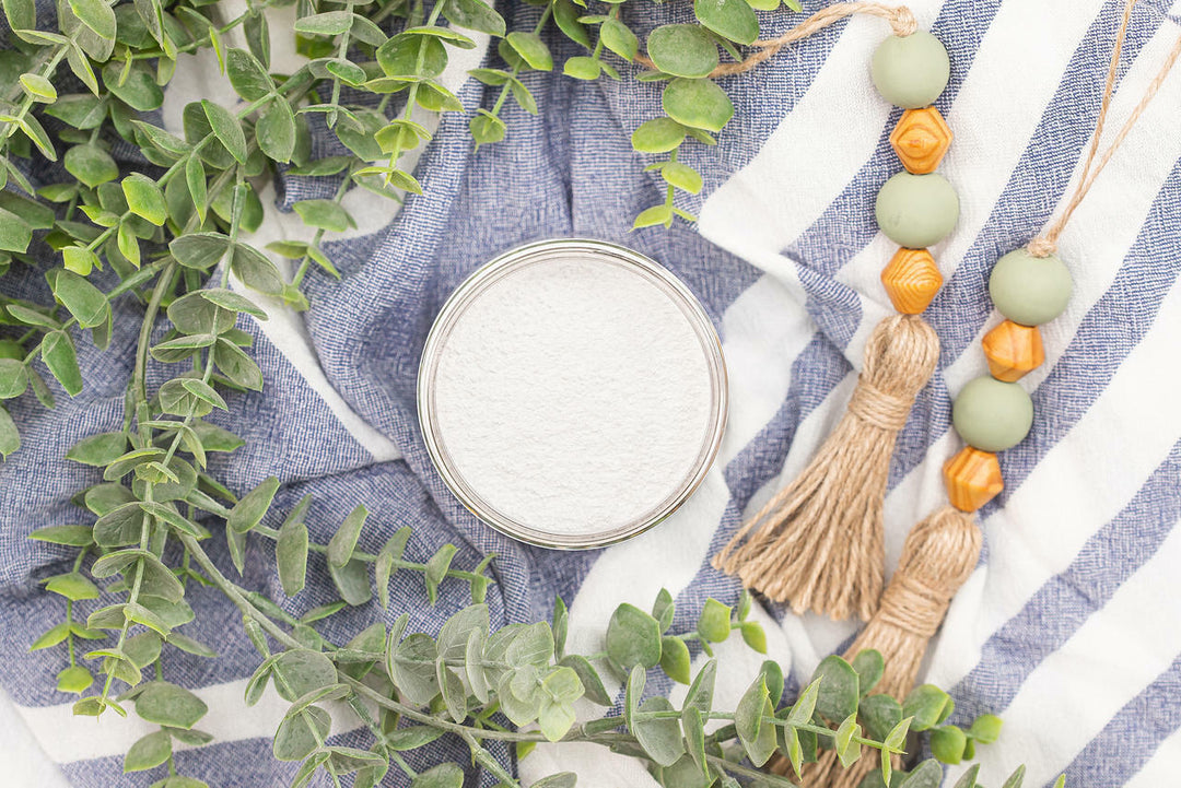 A round silver tin filled with white powder, styled in a flat lay with green eucalyptus leaves, a blue-and-white striped cloth, and a beaded tassel garland.