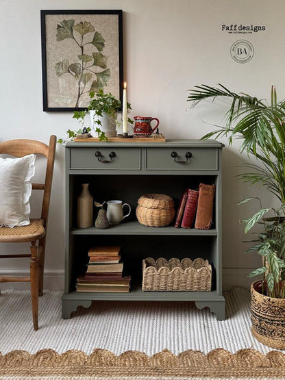 A sage green wooden storage cabinet with two drawers and open shelves, styled in a cozy room setting with a wooden chair, potted plant, and framed botanical print.
