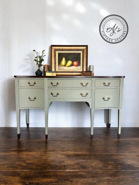 A vintage-style console table painted in a muted sage green, featuring a dark wood top and brass drawer pulls, set against a white wall with a framed still life painting on top.