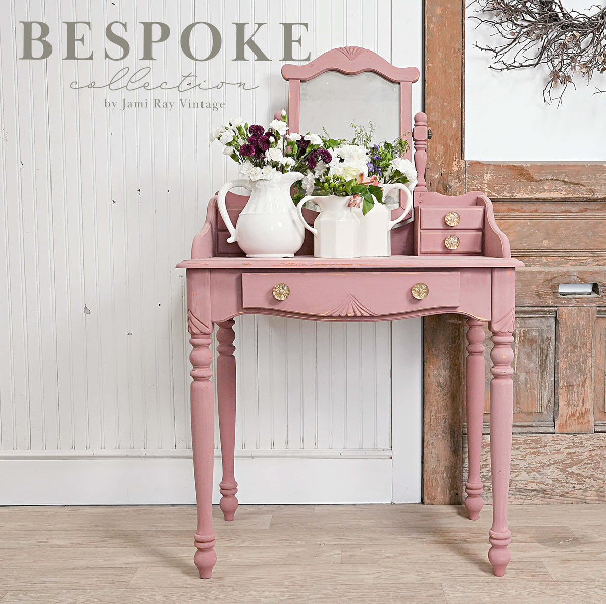 Pink vanity table with mirror and floral arrangements against a wooden wall.