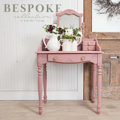Pink vanity table with mirror and floral arrangements against a wooden wall.