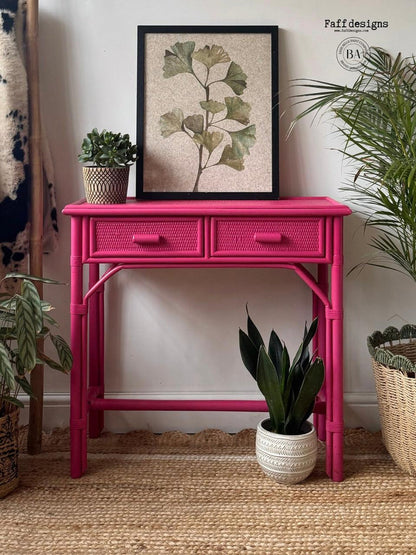 Pink rattan console table with plants and a framed artwork in a room.