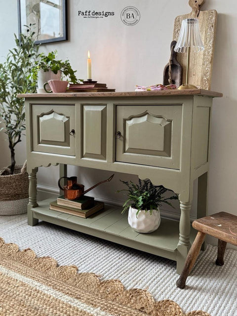 A rustic console table painted in a muted sage green, styled with home decor items, plants, and a small wooden stool in a cozy room setting.