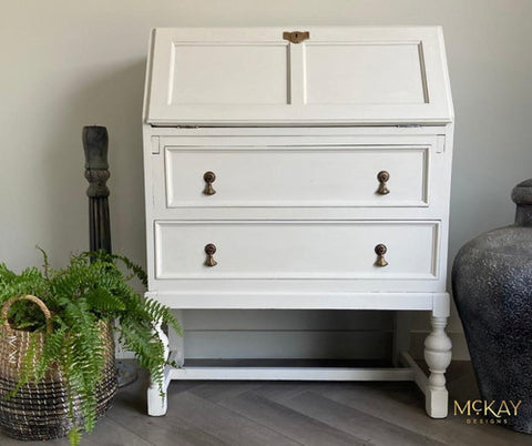 A white painted wooden secretary desk with two drawers and brass knobs, styled in a home setting with a potted fern and a large vase.