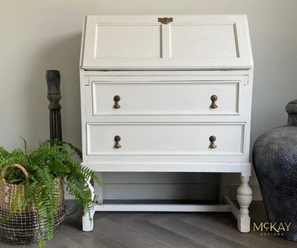 A white painted wooden secretary desk with two drawers and brass knobs, styled in a home setting with a potted fern and a large vase.