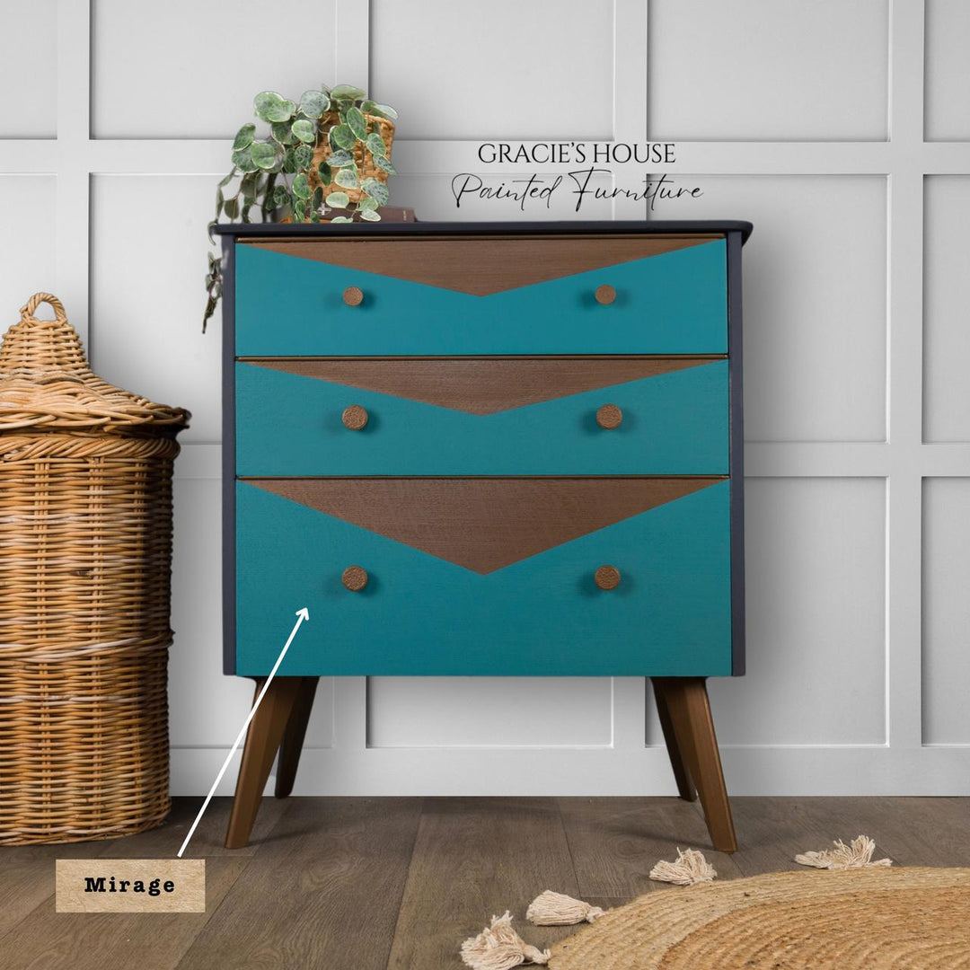 A mid-century modern three-drawer dresser in teal and brown with wooden legs, shown in a styled room setting next to a wicker basket. A text overlay reads 'GRACIE'S HOUSE Painted Furniture' and another label points to the teal drawer front with the text 'Mirage'.