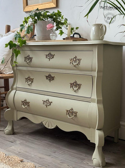A vintage-style three-drawer chest of drawers in a warm off-white/cream finish, decorated with ornate antique bronze drawer pulls and styled with a potted plant and ceramic pitcher on top.