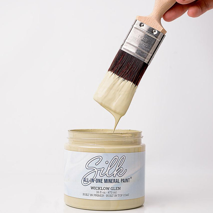 A hand holds a paint brush dipped in thick cream-colored paint above an open jar of Silk All-In-One Mineral Paint in Wicklow Glen.