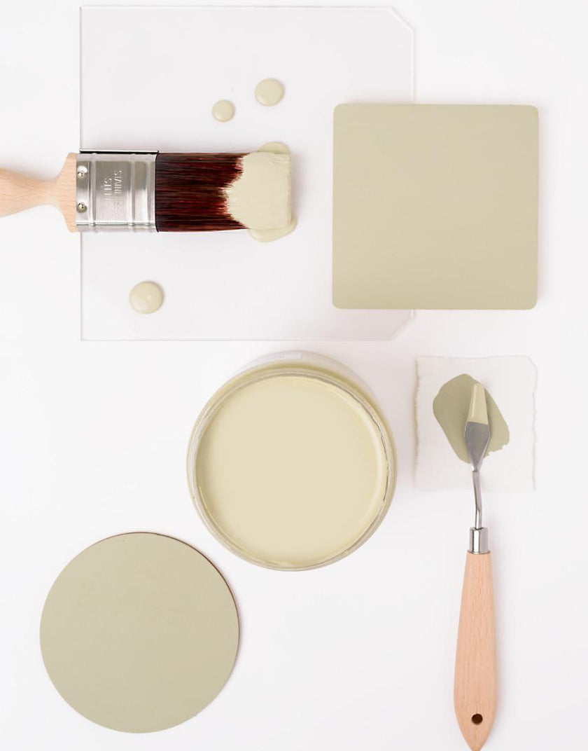 A flat-lay of paint and painting tools in a warm light beige/taupe color, including a paintbrush, a round and square color swatch, a paint can, and a palette knife on a white background.