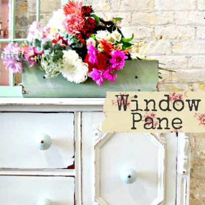 A rustic white wooden cabinet with a distressed finish, decorated with a large floral arrangement in a green box, set against a light brick wall. A beige label overlay reads 'Window Pane'.